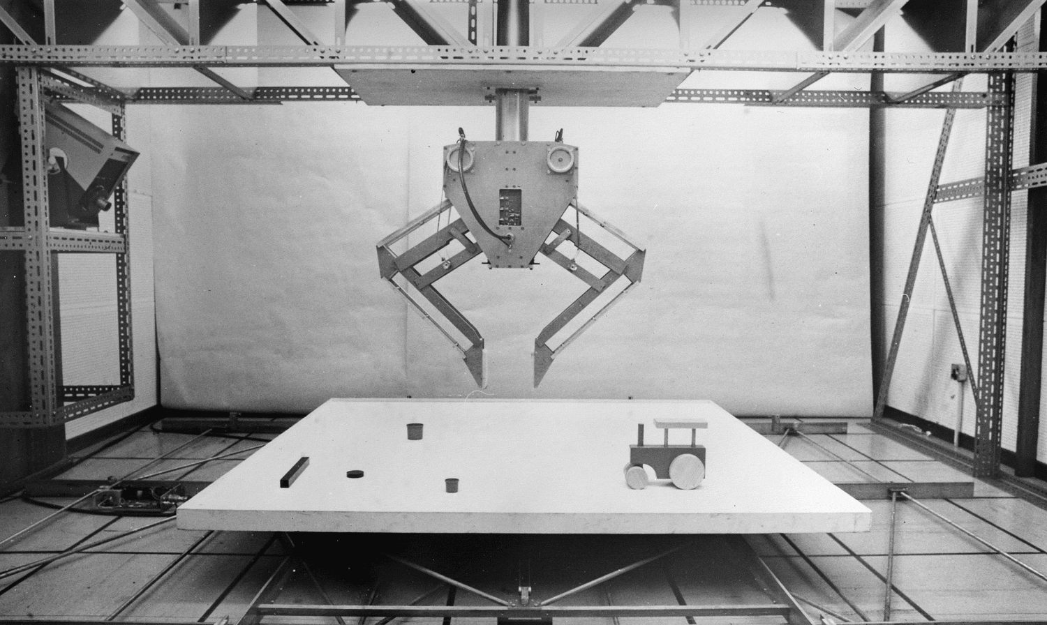 A black and white photograph of a very large metal frame with a metal plate at the top middle. Suspended from the plate is a large metal robot with a triangular section, loosely resembling a face, hanging from a large metal pole, and with two metal arms connected either side. Underneath the robot is a large white square table with some objects on top, including a toy train and some shapes.