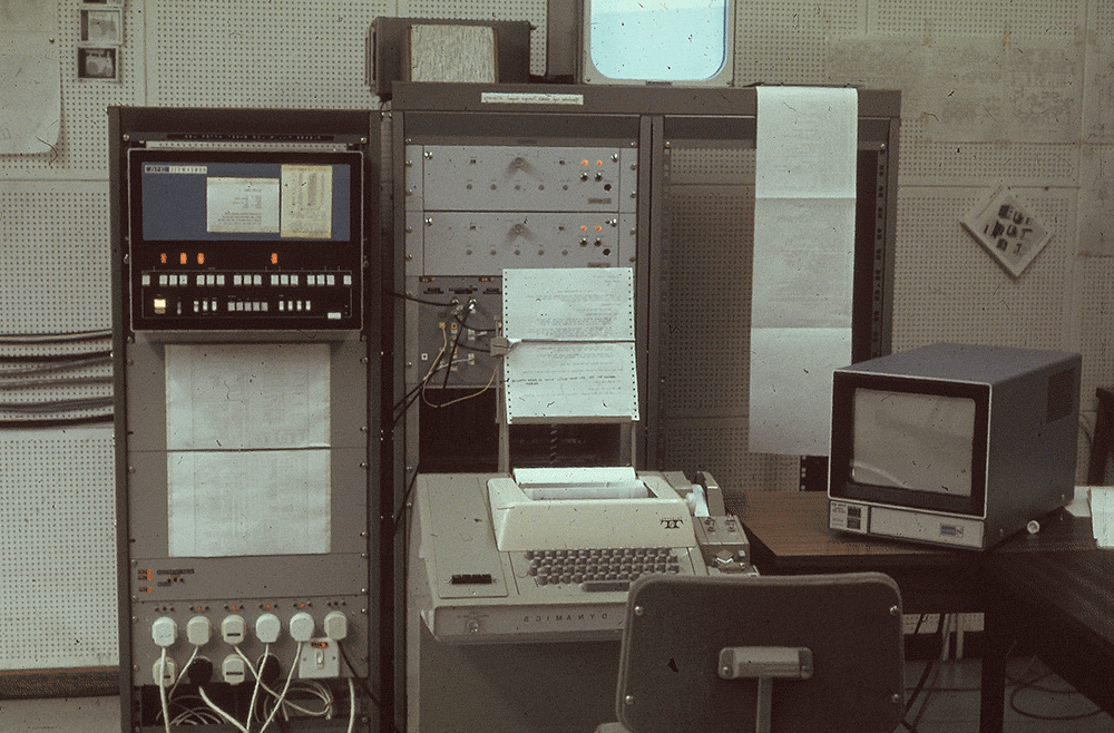 A colour photograph of a 1970s computer room. There are 3 large upright brown metal cases, the size of a fridge, placed next to each other. There are buttons and many plug sockets on the one on the left. On the middle casing, there are wires attached between it and the one on the left. It also has some switches and lights. and a piece of dot matrix paper stuck on the front. There is a roll of dot matrix paper hanging in front of the one on the far right. In front of these casings is a desk and chair with a large tan-coloured typewriting device and a cube-shaped monitor.
