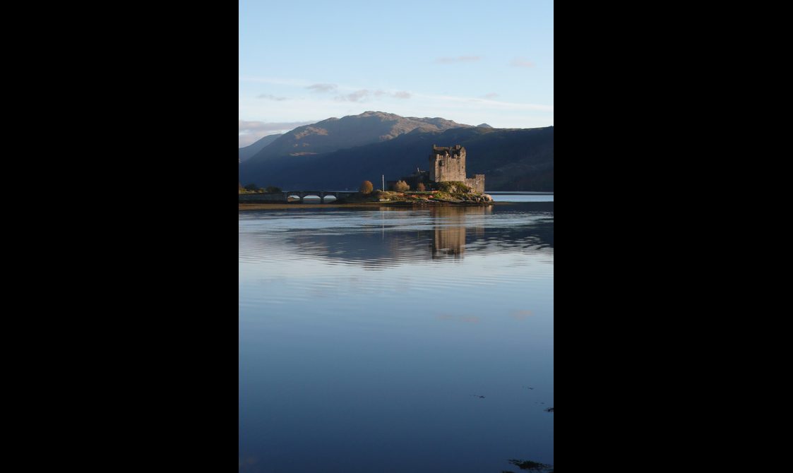 Eilean Donan castle at a slight distance, with the loch in the foreground and hills in the back. It's a clear blue day, and the sky is reflected in the water.