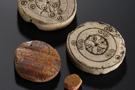 4 circular playing pieces against a black background. 2 are made from pottery, whilst 2 are from bone - and on them has been etched a circular pattern.