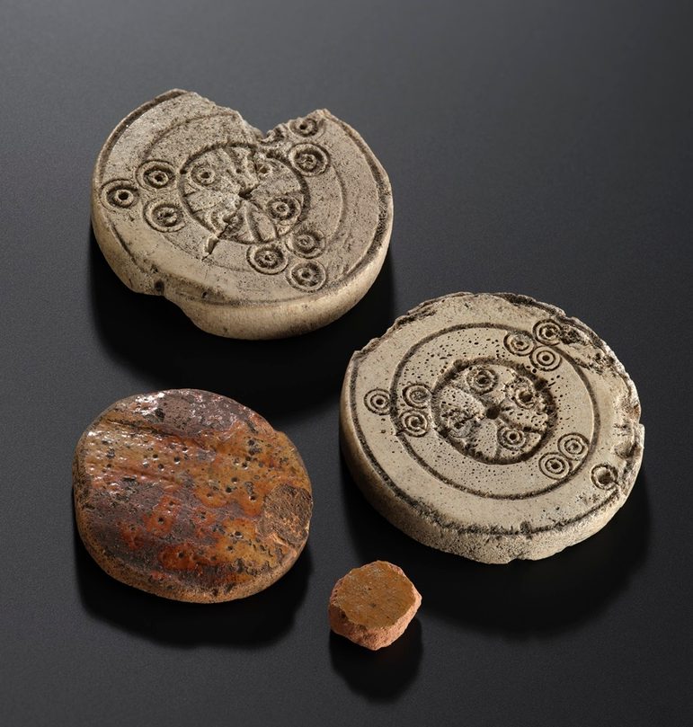 4 circular playing pieces against a black background. 2 are made from pottery, whilst 2 are from bone - and on them has been etched a circular pattern.