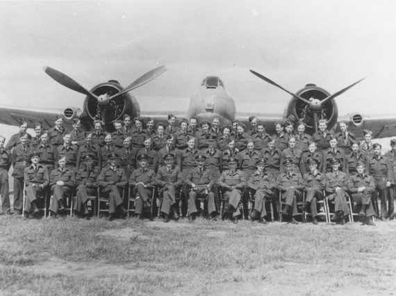 A black and white photograph of pilots in uniform sat and standing in 3 rows, in front of a large propeller plane.