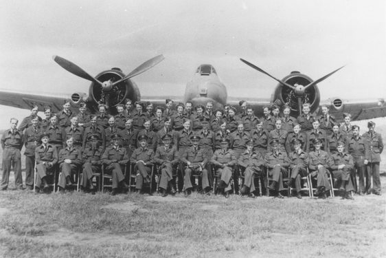 A black and white photograph of pilots in uniform sat and standing in 3 rows, in front of a large propeller plane.