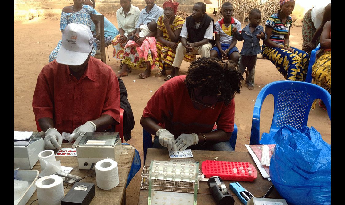 Two scientists sitting at a table testing swabs with a group of patients sitting behind them.