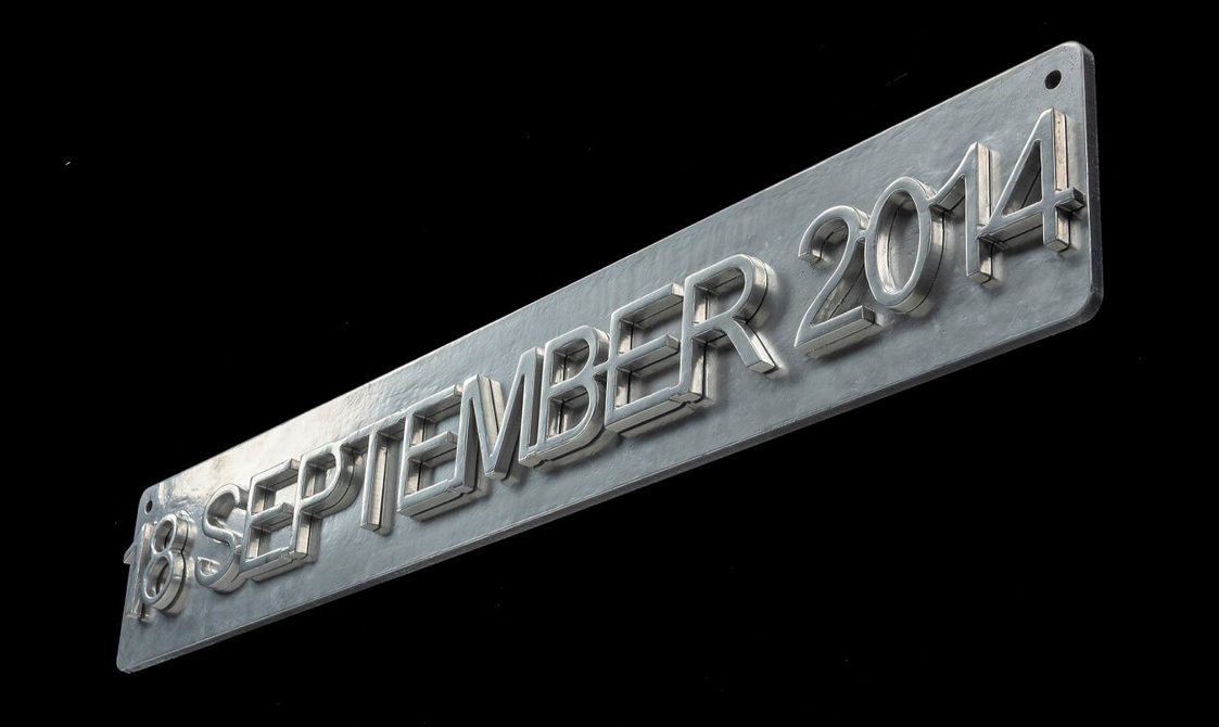 Grey steel sign viewed at a 45 degree low angle, rectangular and narrow with slightly rounded corners. Raised text across it reading '18 SEPTEMBER 2014'.