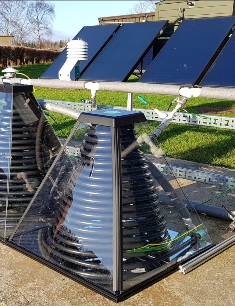 A shiny acrylic pyramid with a black coil sitting on a reflective base. It is sitting outside on pavement in front of some solar panels.