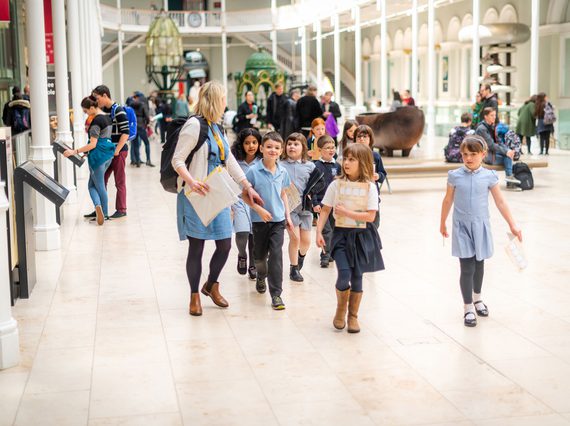 A teacher leads a group of young school children through a large, bright, open gallery.