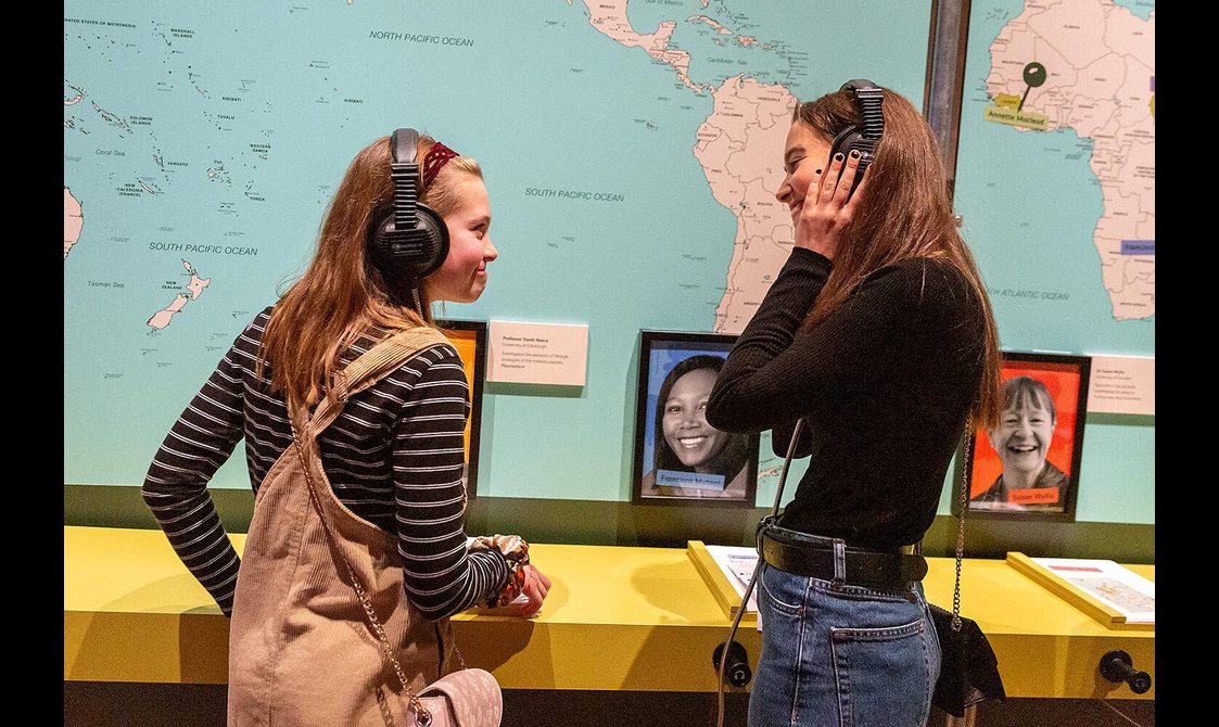 Two visitors listening to people talking through headphones in a museum exhibition.