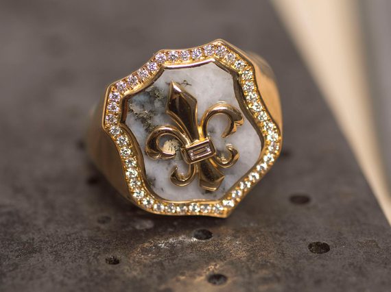 Gold signet ring with a gold fleur de lis on a shield backed by white rock and framed with diamonds.
