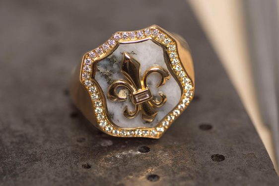 Gold signet ring with a gold fleur de lis on a shield backed by white rock and framed with diamonds.