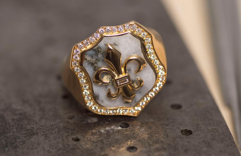 Gold signet ring with a gold fleur de lis on a shield backed by white rock and framed with diamonds.