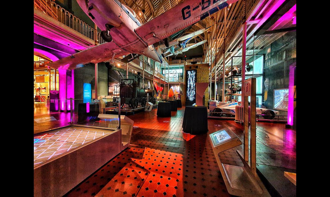 A multi-level museum gallery colourfully lit up for an evening event. There are different aircraft suspended from the ceiling, and different vehicles around the gallery.
