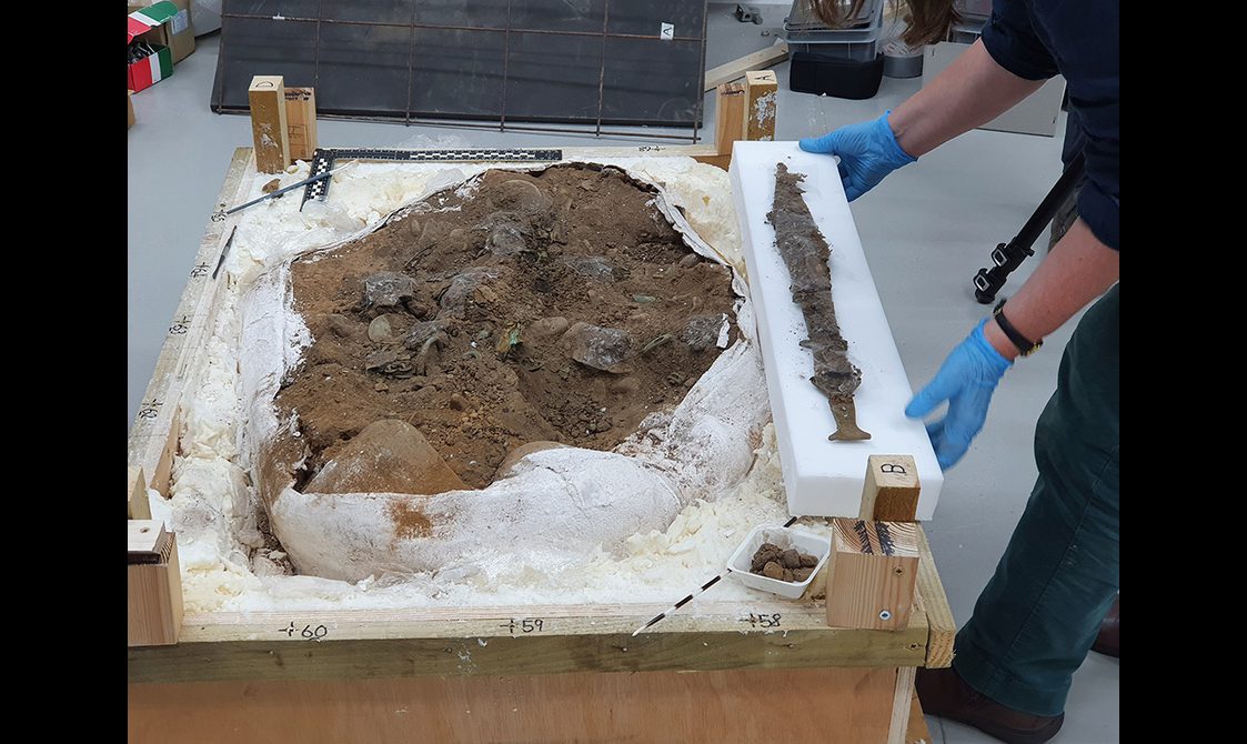 The Peebles Hoard in a 'block' of soil lifted from the ground in the excavation lab. The sword in its scabbard is placed next to it
