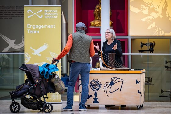A person with a pram is talking to a museum worker, in the grand gallery at the national museum of scotland. The museum worker is at a small stand, with a sign saying 'Engagement team' behind them.