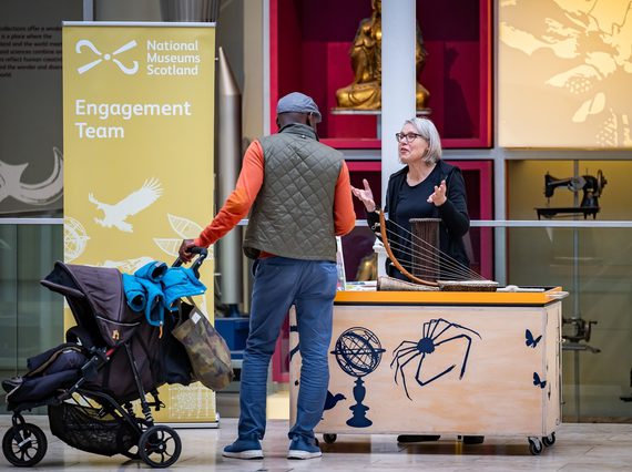 A person with a pram is talking to a museum worker, in the grand gallery at the national museum of scotland. The museum worker is at a small stand, with a sign saying 'Engagement team' behind them.