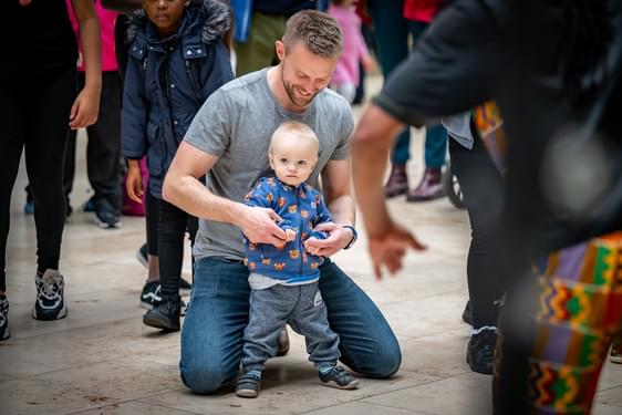 A youg child and his parent or carer are surrounded by a crowd of people and enjoying an event in the grand gallery at the National Museum of Scotland.