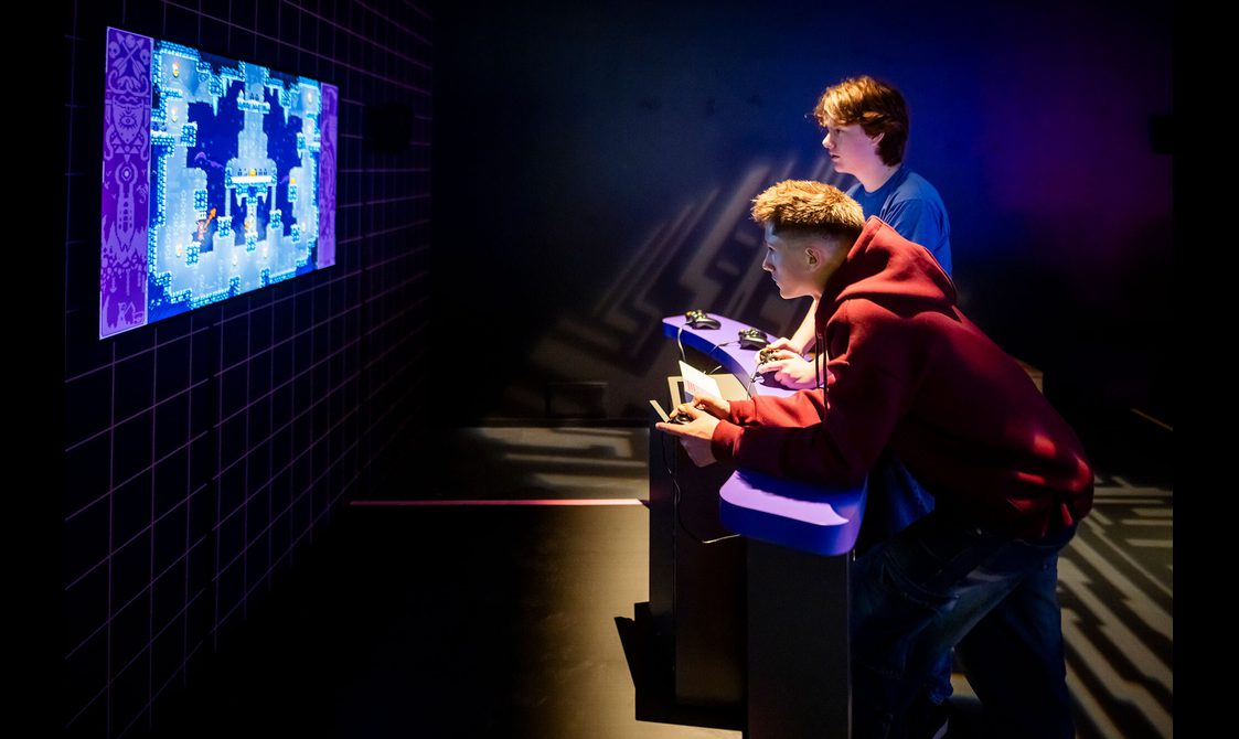 2 visitors to the Game On exhibition are holding consoles and playing bomberman.
