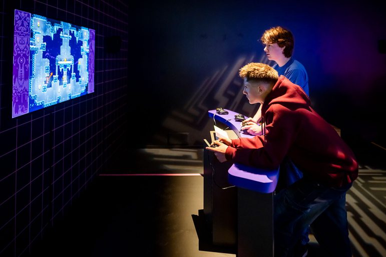 2 visitors to the Game On exhibition are holding consoles and playing bomberman.