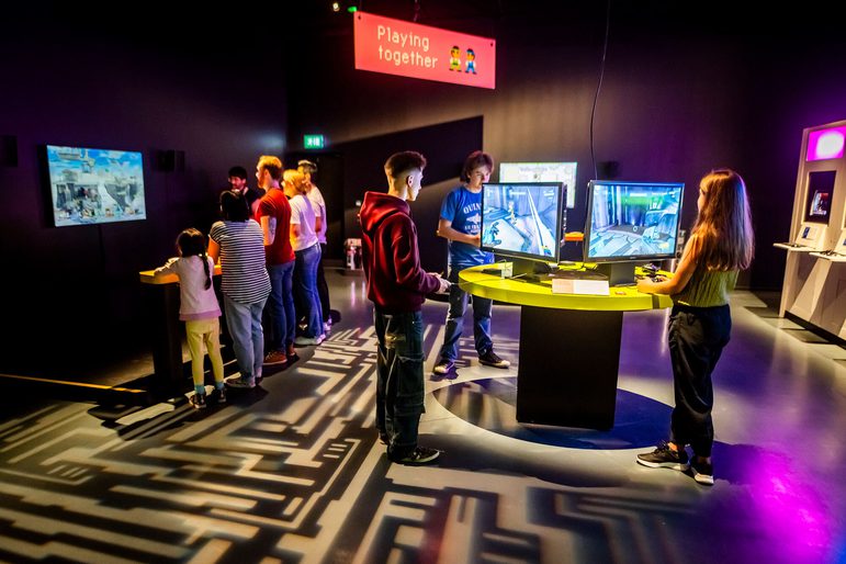 A group of visitors are dispersed across the Game On exhibition gallery, each playing a different video game.