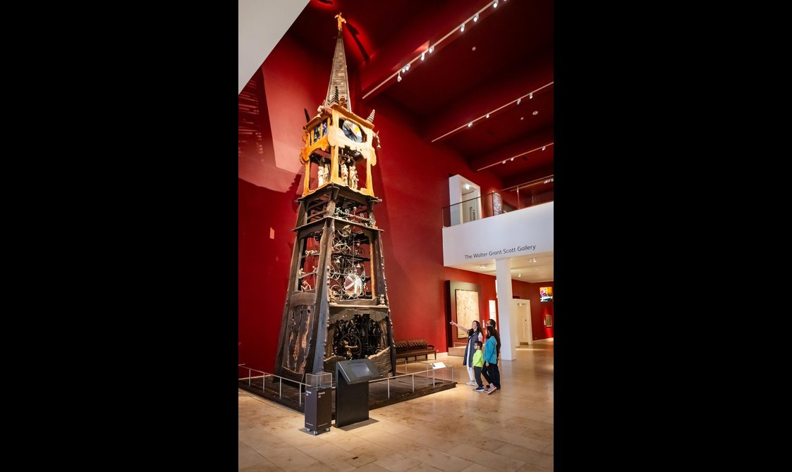 A family of four in a museum looking at a large ornate wooden clock with carved wooden models.