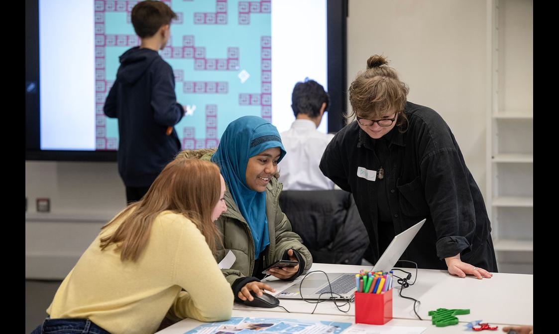 A large display screen in the background shows a grid of a computer game whilst in the foreground two smiling adults look at the laptop screen in front of a teenager who is smiling.