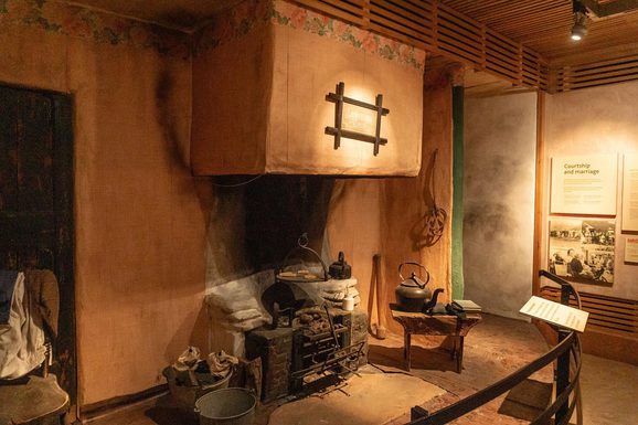 A museum display of an old farmhouse kitchen with a chimney and stove surrounded by wooden stools.