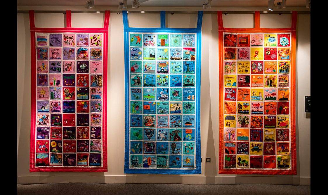 Three large vibrant quilts hanging against a yellow wall. Made up of colourful squares showing patterns, shapes and words of hope for the future.