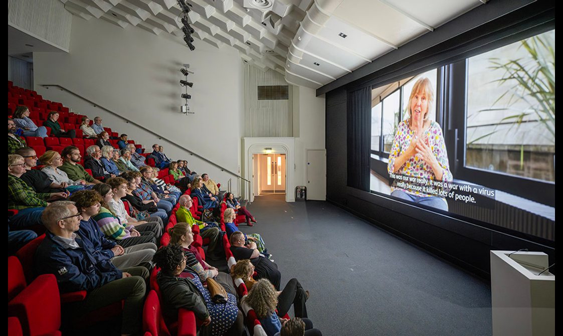 Group of people in auditorium watching Injecting Hope: The Deaf Experience film on large screen.
