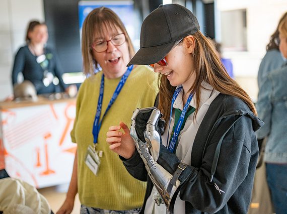 A young person in glasses and a hat looking at a robotic hand.