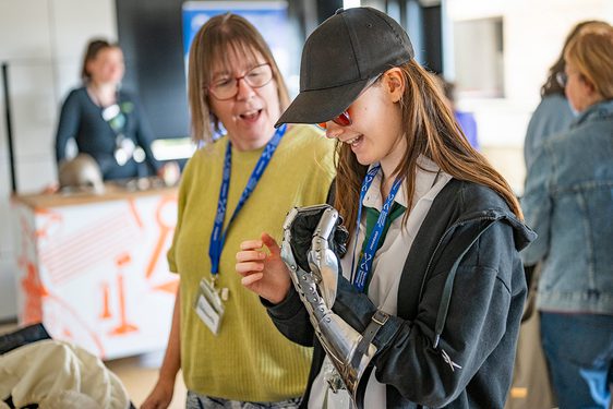 A young person in glasses and a hat looking at a robotic hand.