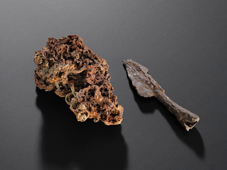 a lump of slightly rust coloured rock is on the left, with tiny circular pieces of interlocking metal just visible. Next to it, on the right, is a slightly broken arrowhead.