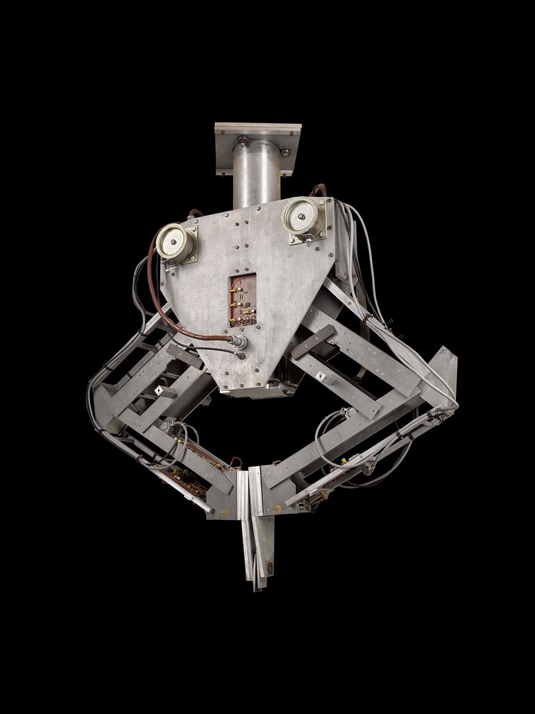 A colour photograph of a metal robotic machine against a black background. The machine is has a mounting brace at the top and a triangular central section which loosely resembles a face. The central section has two mechanical arms attached either side, bents ate the 'elbow', and with flat clasps resembling hands at the bottom of each. The 'hands' are touching in the centre, creating an inverted triangle with the arms.