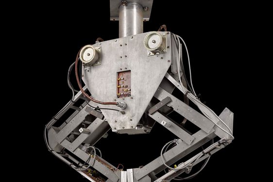 A colour photograph of a metal robotic machine against a black background. The machine is has a mounting brace at the top and a triangular central section which loosely resembles a face. The central section has two mechanical arms attached either side, bents ate the 'elbow', and with flat clasps resembling hands at the bottom of each. The 'hands' are touching in the centre, creating an inverted triangle with the arms.