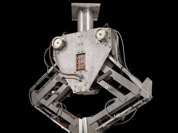 A colour photograph of a metal robotic machine against a black background. The machine is has a mounting brace at the top and a triangular central section which loosely resembles a face. The central section has two mechanical arms attached either side, bents ate the 'elbow', and with flat clasps resembling hands at the bottom of each. The 'hands' are touching in the centre, creating an inverted triangle with the arms.