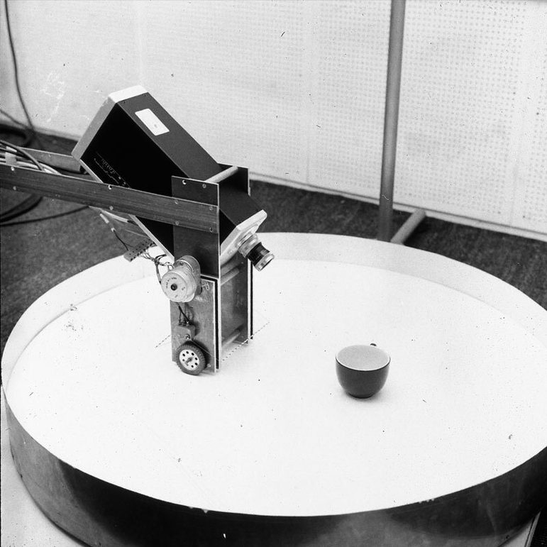 A black and white photograph id a circular table with a coffee cup on top. Next to that, to the left, is a black metal arm with a camera mounted on the end, and a silver vertical metal box with very small wheels. The wheels are on top of the table.