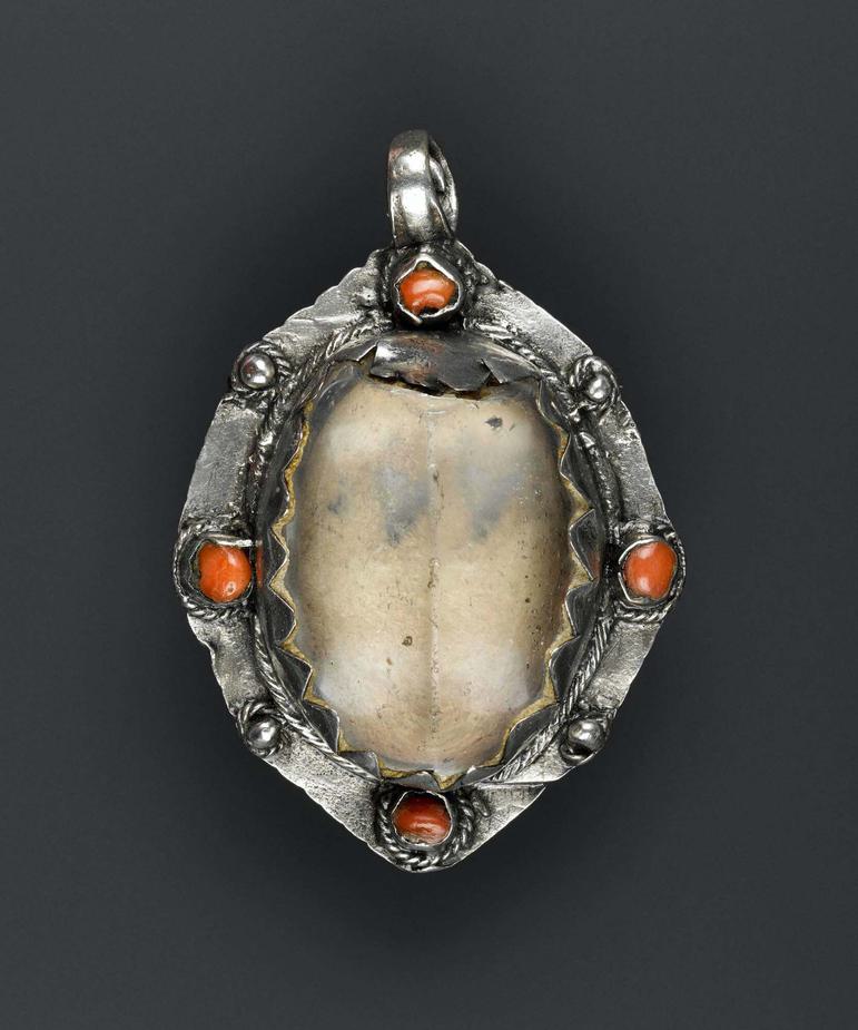 An oval silver pendant with a crystal setting. One side of the crystal is damaged. The mount is decorated with four pieces of red coral alternating with four silver balls.