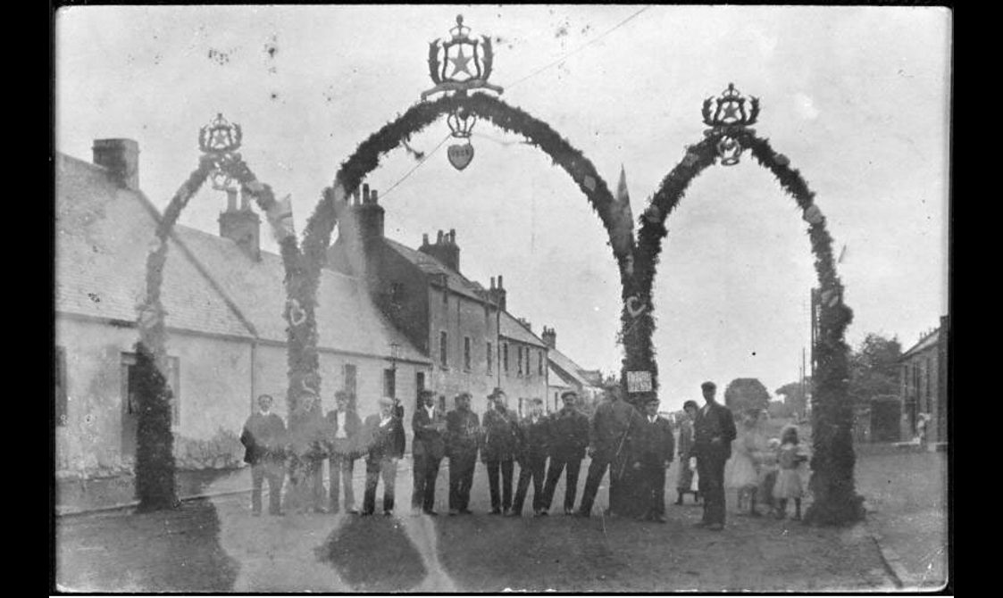 A group of people standing under huge arches built in the middle of the street. The highest point of the arches is nearly as high as the roof of the cottages on the street.