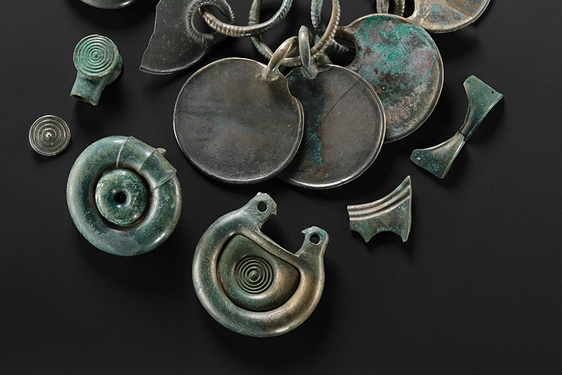 Twelve bronze objects, including some rounded fittings with concentric circle designs and a rattle pendant. They are all silvery, with some green corrosion on the surface.