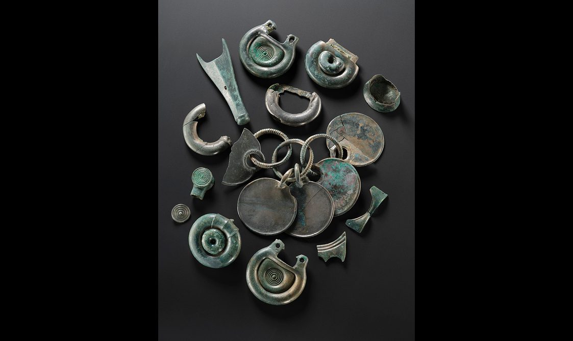 Twelve bronze objects, including some rounded fittings with concentric circle designs and a rattle pendant. They are all silvery, with some green corrosion on the surface.