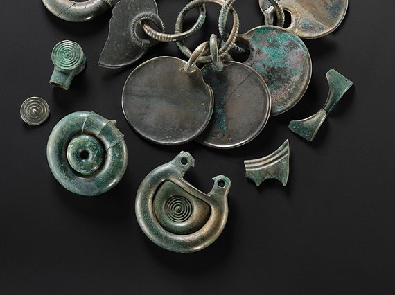 Twelve bronze objects, including some rounded fittings with concentric circle designs and a rattle pendant. They are all silvery, with some green corrosion on the surface.