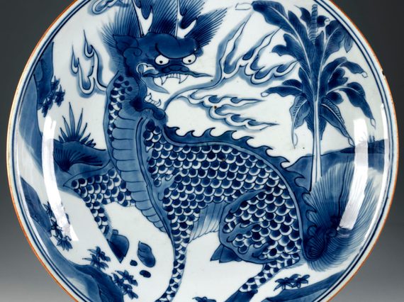 Circular plate with a blue dragon figure in the center wit ha palm tree and landscape scenery in the background.