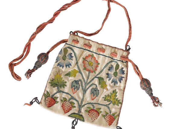 Square shaped purse decorated with blue and red flowers at the top, and six strawberries at the bottom. A red string attached to the top of thee purse, and three black tassels hang at the bottom of the purse.
