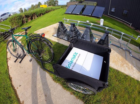 A bike in a stand on a pavement. It has a black trailer attached with a white box in it that says 'SolarisKit' in green. Behind the bike there are some pyramid shaped devices with coils inside, and there are some solar panels in the far background.