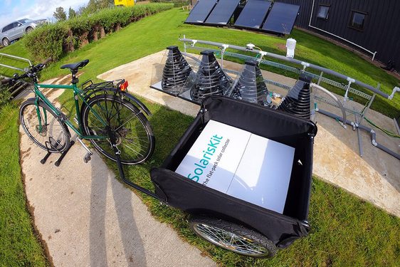 A bike in a stand on a pavement. It has a black trailer attached with a white box in it that says 'SolarisKit' in green. Behind the bike there are some pyramid shaped devices with coils inside, and there are some solar panels in the far background.