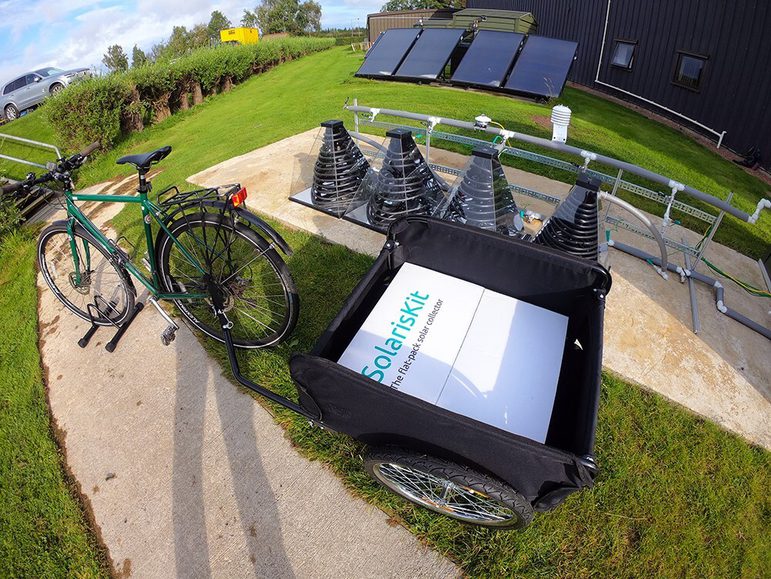 A bike in a stand on a pavement. It has a black trailer attached with a white box in it that says 'SolarisKit' in green. Behind the bike there are some pyramid shaped devices with coils inside, and there are some solar panels in the far background.