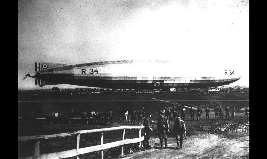 A black and white photograph of a long blimp-like aircraft in an airfield with several men watching.