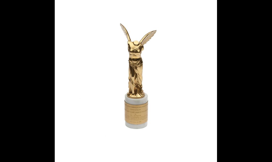 A gold trophy of the body of a woman wearing a long dress with wings standing on a round stand with a gold plaque with an inscription