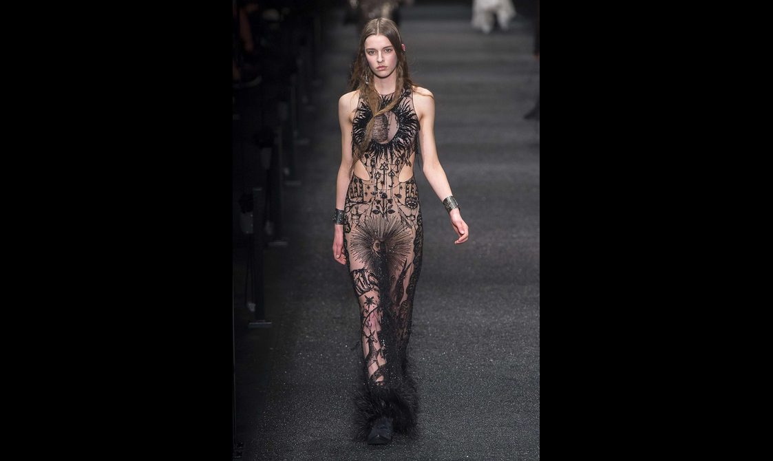 A model walking a runway wearing a black sheer dress with suns, flowers, and animals embroidered on it.