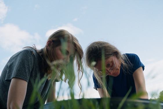 2 women are looking down at something on the ground. The framing of the shot looks like the photo has been taken from grass level in a field, and the sky is blue behind them.