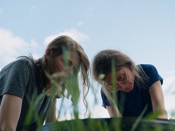 2 women are looking down at something on the ground. The framing of the shot looks like the photo has been taken from grass level in a field, and the sky is blue behind them.